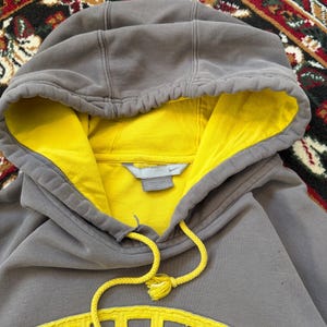 May include: A gray Nike hoodie with a yellow interior and a yellow Nike logo on the front. The hoodie has a drawstring and a hood. The image is a close-up shot of the hoodie.