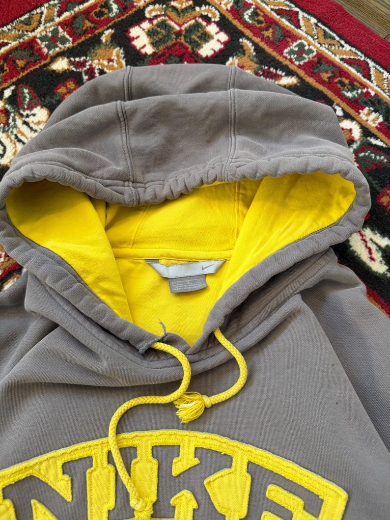 May include: A gray Nike hoodie with a yellow interior and a yellow Nike logo on the front. The hoodie has a drawstring and a hood. The image is a close-up shot of the hoodie.