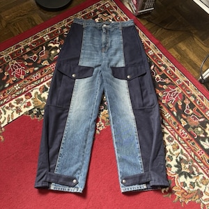 May include: High-waisted cargo pants featuring a color-blocked design with a light blue denim center panel and dark navy blue side panels. The pants include two large flap pockets on the thighs, a button fly closure, and adjustable straps at the cuffs.