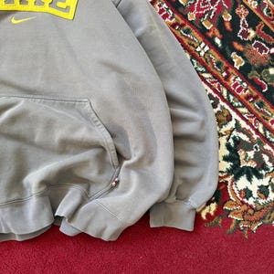 May include: A gray Nike hoodie with a yellow logo and a front pocket. The word "NIKE" is displayed in yellow block letters with a yellow Nike swoosh. The hoodie has a drawstring hood and ribbed cuffs.