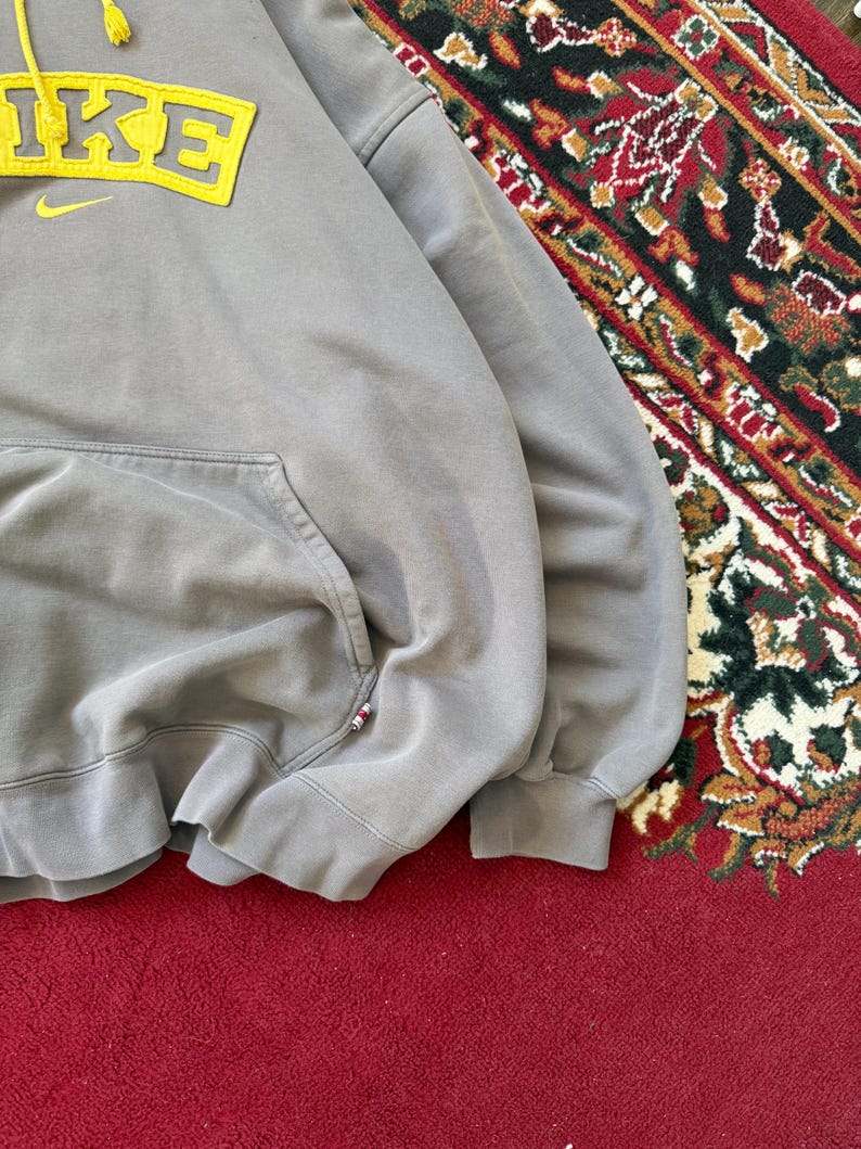 May include: A gray Nike hoodie with a yellow logo and a front pocket. The word "NIKE" is displayed in yellow block letters with a yellow Nike swoosh. The hoodie has a drawstring hood and ribbed cuffs.