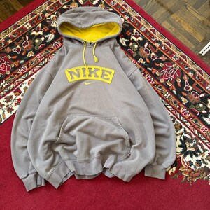 May include: A gray Nike hoodie with a yellow-lined hood and a large yellow Nike logo across the chest. The hoodie has a front pocket and yellow drawstrings. The garment is displayed on a red patterned rug.