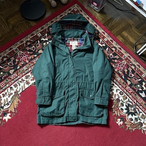 Puede incluir: Chaqueta con capucha verde oscuro con cierre de botones y múltiples bolsillos. La chaqueta tiene un forro de cuadros visible en la capucha y el cuello. La chaqueta está sobre una alfombra roja estampada.