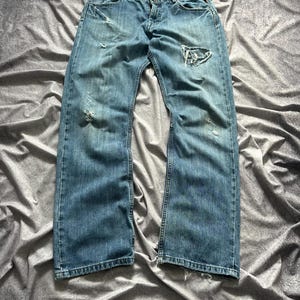 May include: Light blue denim jeans with distressed details, including rips and fading. The jeans have a classic five-pocket design and a relaxed fit. The fabric appears worn and softened, suggesting a vintage or well-loved aesthetic.