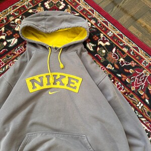 May include: Gray Nike hoodie with a yellow interior hood and drawstring. The word "NIKE" is displayed in yellow across the chest, with a small Nike swoosh logo below. The hoodie has a front pocket.