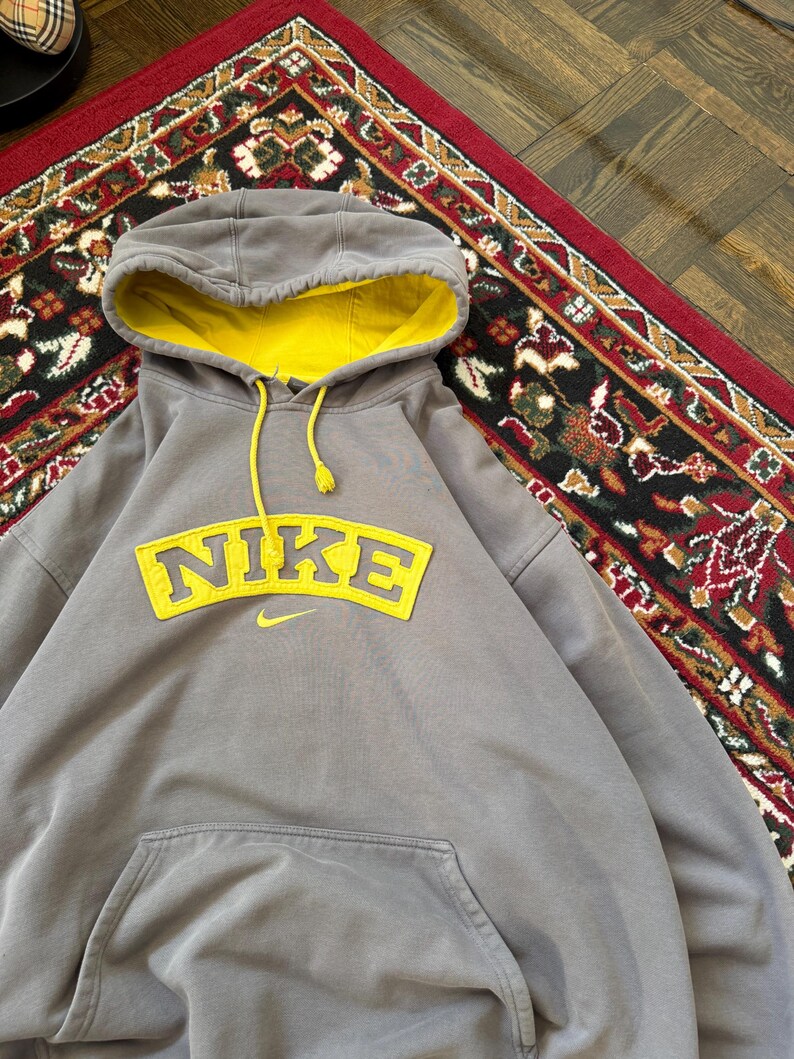 May include: Gray Nike hoodie with a yellow interior hood and drawstring. The word "NIKE" is displayed in yellow across the chest, with a small Nike swoosh logo below. The hoodie has a front pocket.