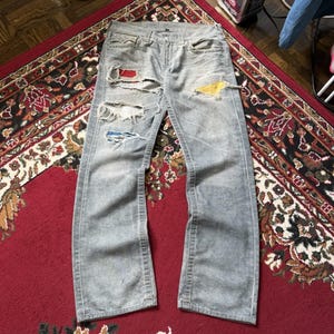 Könnte beinhalten: Hellgraue Jeans aus Denim im Used-Look mit mehreren Rissen und Löchern. Die ausgefransten Stellen geben den Blick auf darunterliegende Stoffflicken in Rot, Gelb und Blau frei. Die Jeans verfügt über eine Knopfleiste, Gürtelschlaufen und fünf Taschen.