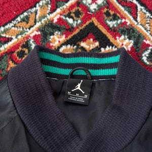 Może przedstawiać: Czarna kurtka bomberka z prążkowanym kołnierzem w zielono-czarne paski. Kurtka posiada czarną metkę z logo Jumpman i rozmiarem "M". Na metce widnieje r&oacute;wnież napis "MADE IN CHINA".