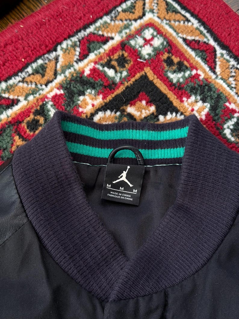 Może przedstawiać: Czarna kurtka bomberka z prążkowanym kołnierzem w zielono-czarne paski. Kurtka posiada czarną metkę z logo Jumpman i rozmiarem "M". Na metce widnieje r&oacute;wnież napis "MADE IN CHINA".
