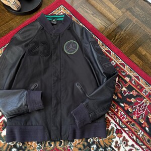 Może przedstawiać: Czarna kurtka bomberka z numerem "23" z przodu i okrągłą naszywką z logo "Jordan". Kurtka ma prążkowany kołnierz, mankiety i d&oacute;ł. Kurtka jest prezentowana na czerwonym dywanie.