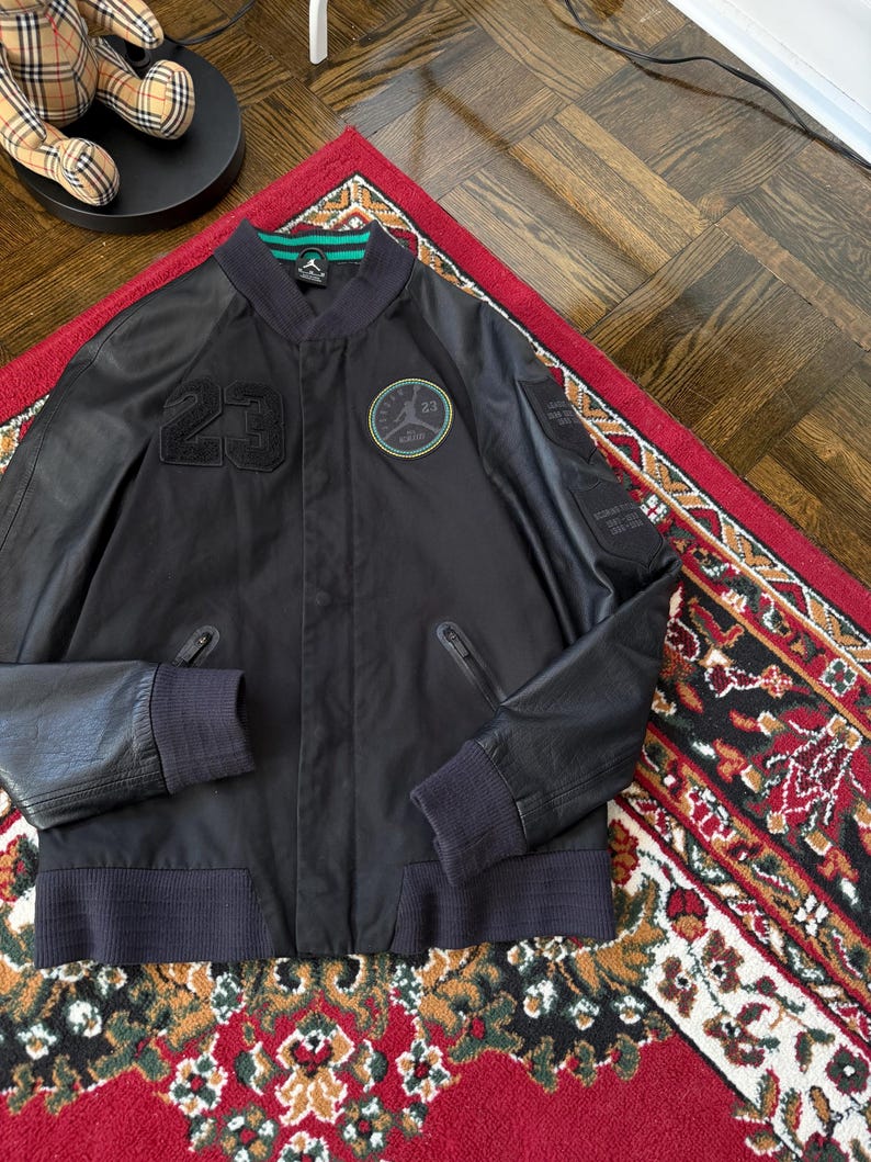 Może przedstawiać: Czarna kurtka bomberka z numerem "23" z przodu i okrągłą naszywką z logo "Jordan". Kurtka ma prążkowany kołnierz, mankiety i d&oacute;ł. Kurtka jest prezentowana na czerwonym dywanie.