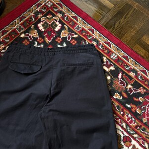 May include: Navy blue trousers with a drawstring waist and two back pockets. The trousers are made of a smooth fabric and are displayed on a patterned rug with red, black, and gold accents. The trousers are casual.