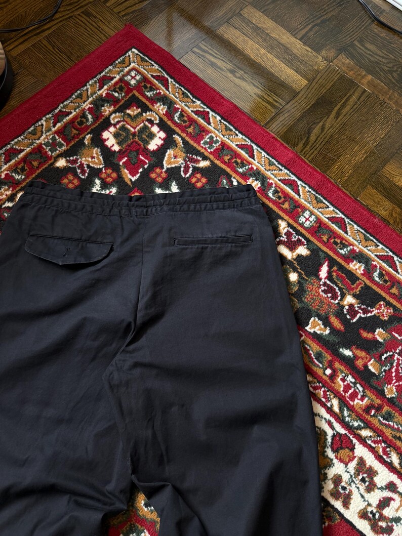 May include: Navy blue trousers with a drawstring waist and two back pockets. The trousers are made of a smooth fabric and are displayed on a patterned rug with red, black, and gold accents. The trousers are casual.