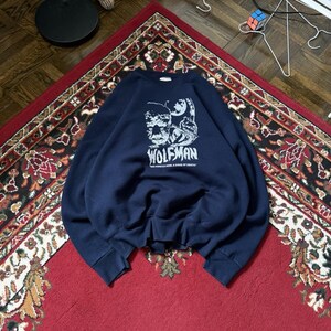 May include: A navy blue sweatshirt with a graphic of the Wolfman, the text "WOLFMAN" and "THE WEREWOLF'S CURSE OF DEATH". The sweatshirt is on a red patterned rug.