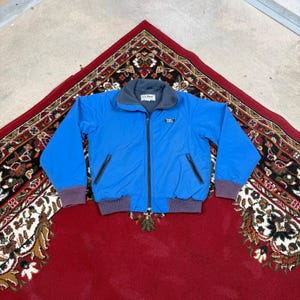 May include: A bright blue zip-up jacket featuring a dark gray fleece-lined collar, dark gray ribbed cuffs and hem, and two zippered side pockets. The L.L. Bean brand logo is visible on the chest and on the interior neck label.