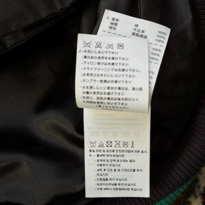 Może przedstawiać: Zbliżenie czarnej kurtki z zielono-białym paskiem. Dołączone są dwie białe metki z japońskim tekstem i symbolami pielęgnacji. Etykiety zawierają skład tkaniny i instrukcje prania.