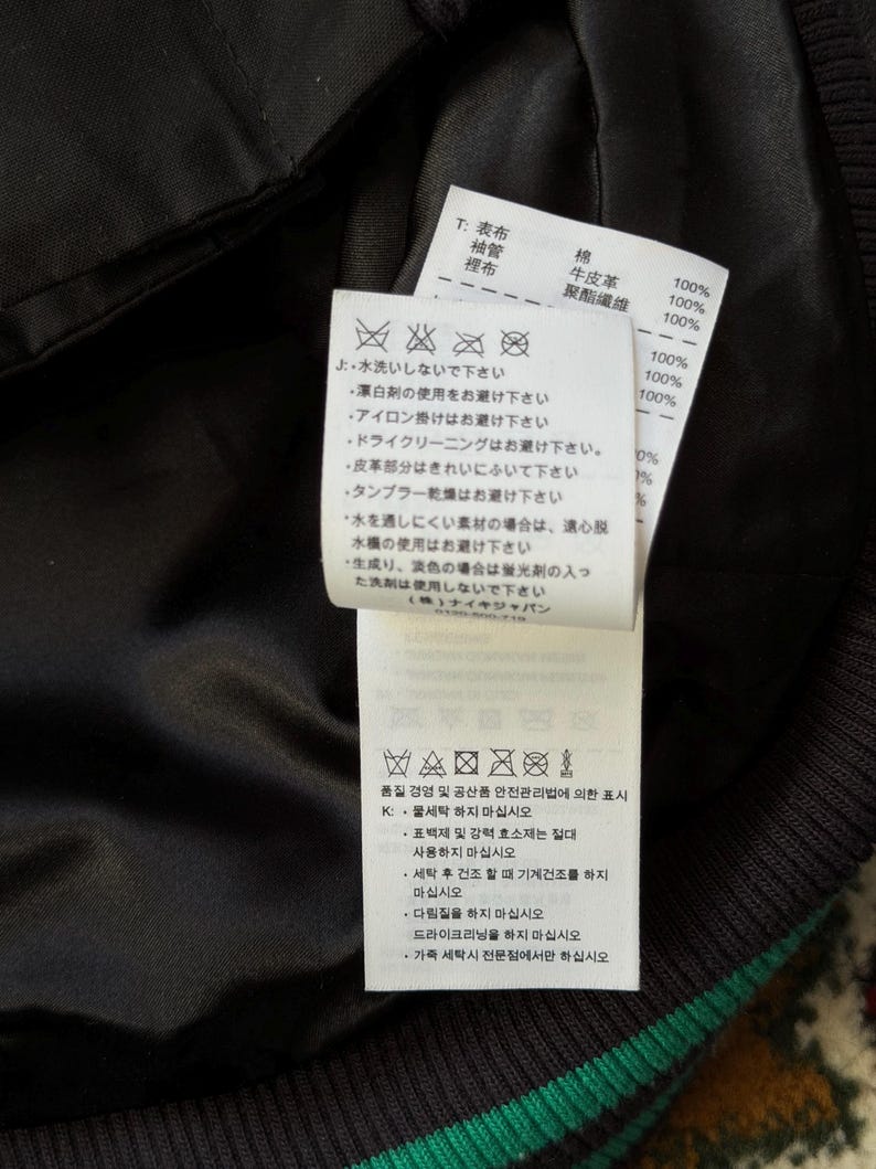 Może przedstawiać: Zbliżenie czarnej kurtki z zielono-białym paskiem. Dołączone są dwie białe metki z japońskim tekstem i symbolami pielęgnacji. Etykiety zawierają skład tkaniny i instrukcje prania.