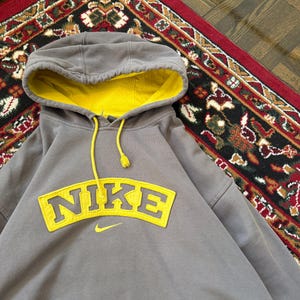May include: A gray Nike hoodie with a yellow interior hood and a yellow Nike logo on the front. The hoodie has a drawstring and a kangaroo pocket. The hoodie is made of a soft, comfortable material.