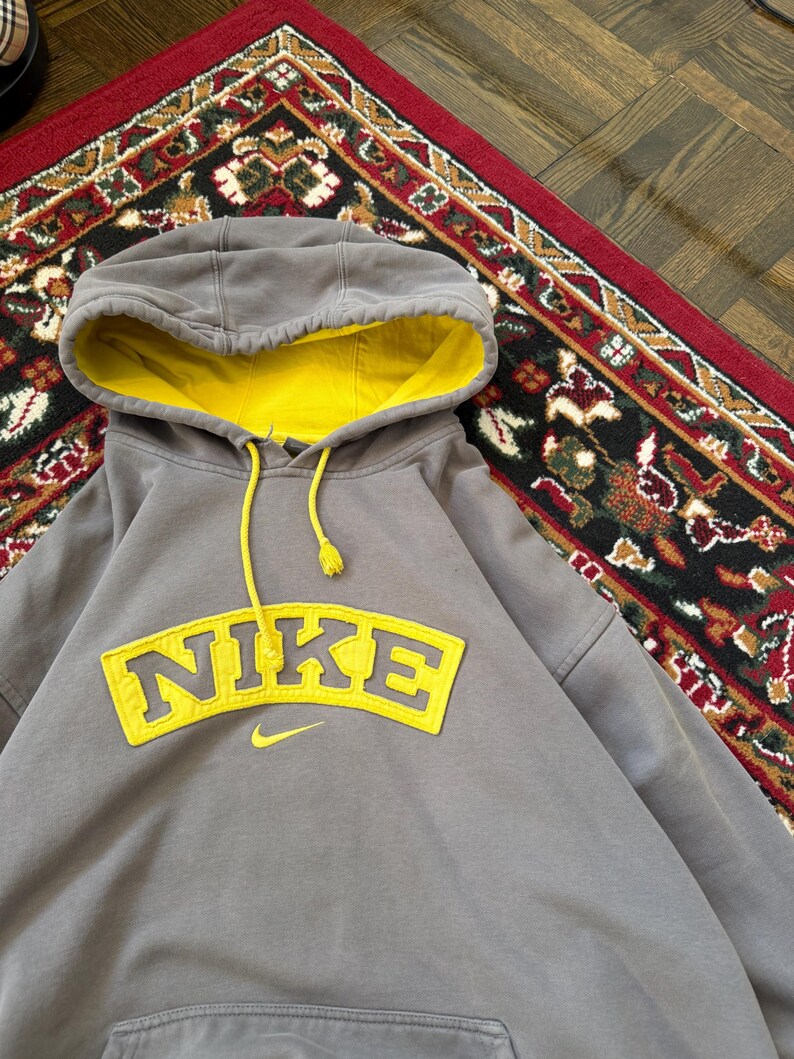 May include: A gray Nike hoodie with a yellow interior hood and a yellow Nike logo on the front. The hoodie has a drawstring and a kangaroo pocket. The hoodie is made of a soft, comfortable material.