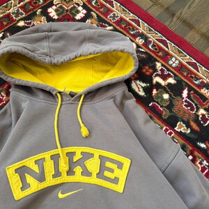 May include: Gray Nike hoodie with a yellow interior and drawstring. The word "NIKE" is prominently displayed in yellow across the chest. A small yellow Nike swoosh logo is below the word. The hoodie is on a patterned rug.