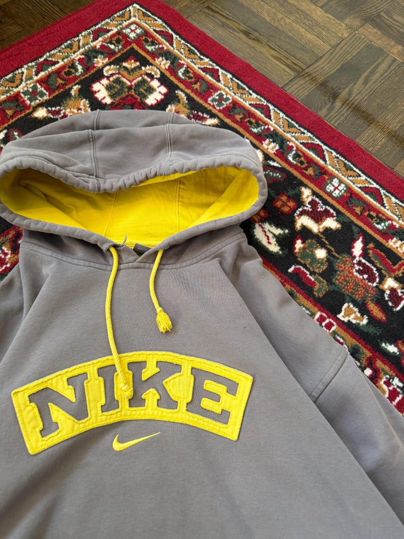 May include: Gray Nike hoodie with a yellow interior and drawstring. The word "NIKE" is prominently displayed in yellow across the chest. A small yellow Nike swoosh logo is below the word. The hoodie is on a patterned rug.