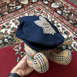 Puede incluir: Gorra de estilo newsboy azul marino con un emblema de hoja de arce que dice "TORONTO MAPLE LEAFS". La gorra está sobre un oso de peluche con un estampado de cuadros. El fondo es una alfombra estampada.