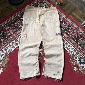 May include: Beige Carhartt work trousers featuring a straight-leg fit, two back pockets, and utility loops. The trousers show signs of wear, including dark stains, scuffs, and frayed hems at the bottom of both legs. A small rectangular brand logo is visible.