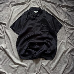 May include: Black short-sleeve button-down shirt with a textured pattern. The shirt has a classic collar, a chest pocket, and a button-up front. The fabric appears to be a lightweight material.