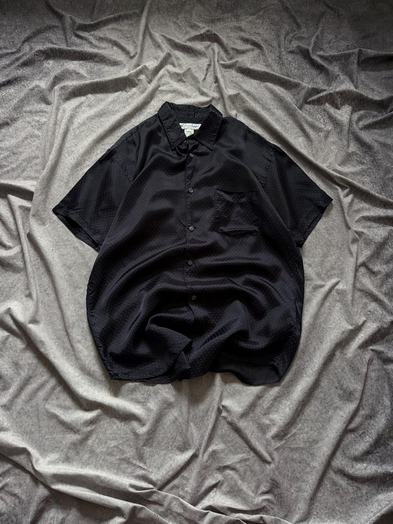 May include: Black short-sleeve button-down shirt with a textured pattern. The shirt has a classic collar, a chest pocket, and a button-up front. The fabric appears to be a lightweight material.
