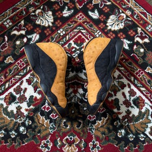 Op de afbeelding: Paar zwarte en bruine sneakers met een bovenwerk van getextureerd leer. De schoenen staan op een kleed met patronen in rood, zwart en cr&egrave;me. De sneakers hebben een high-top design.