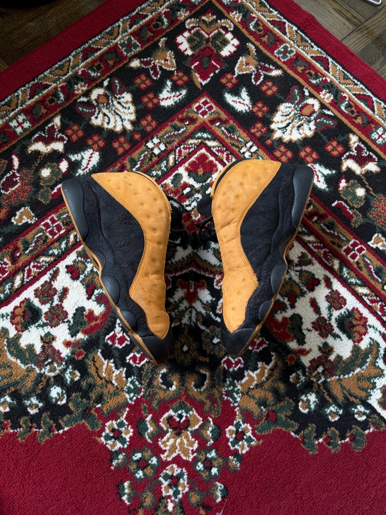 Op de afbeelding: Paar zwarte en bruine sneakers met een bovenwerk van getextureerd leer. De schoenen staan op een kleed met patronen in rood, zwart en cr&egrave;me. De sneakers hebben een high-top design.