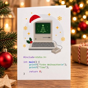 Könnte beinhalten: Eine weiße Grußkarte mit einem alten Computer, der eine rote Weihnachtsmütze trägt und einen pixeligen Weihnachtsbaum auf dem Bildschirm zeigt. Darunter steht der Code #include <stdio.h>, int main() { printf("Frohe Weihnachten
"); printf("Timo"); return 0; }.