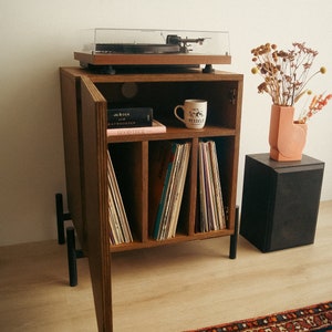 Peut inclure: Un meuble en bois pour tourne-disque avec un tourne-disque sur le dessus, une étagère avec des disques, des livres et une tasse, et une enceinte noire sur le sol. Le meuble a une porte ouverte, révélant les étagères à l'intérieur. Le meuble est sur un sol en bois avec un tapis rouge et bleu devant.