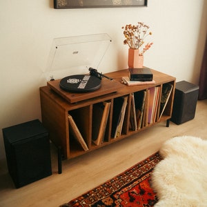 May include: A wooden record player with a clear cover sits atop a wooden cabinet filled with vinyl records. A framed portrait hangs above the cabinet. Two black speakers flank the cabinet, and a patterned rug is in front.