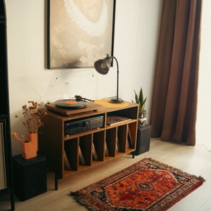 May include: A wooden record player stand with six shelves for vinyl records. The stand has a turntable on top and a stereo receiver below. A lamp is on the left side of the stand, and a potted plant is on the right. A red and black patterned rug is on the floor in front of the stand.