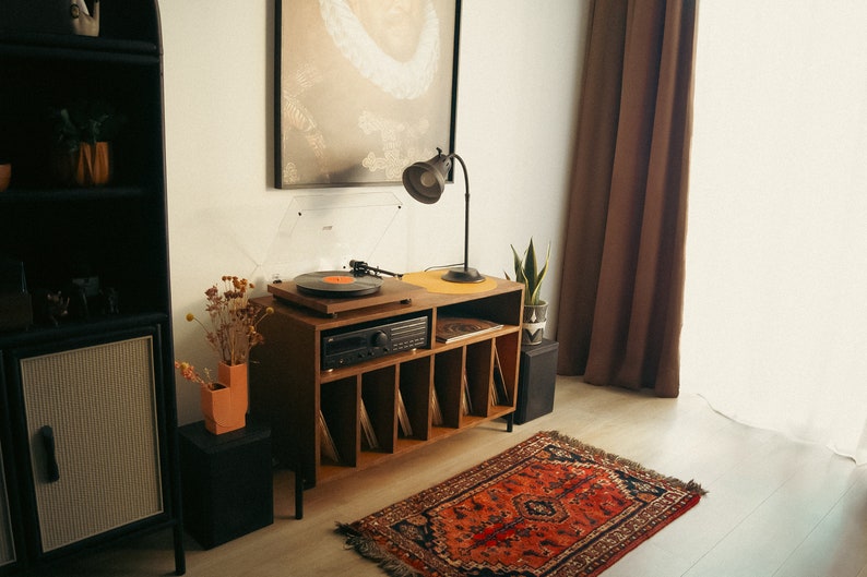 May include: A wooden record player stand with six shelves for vinyl records. The stand has a turntable on top and a stereo receiver below. A lamp is on the left side of the stand, and a potted plant is on the right. A red and black patterned rug is on the floor in front of the stand.