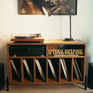 May include: A wooden record player stand with a turntable, amplifier, and shelves for vinyl records. The stand has a black lamp with a white shade and a plant in a black and white pot on top. The text "OPTIONAL BACKPANEL" is printed on the stand.