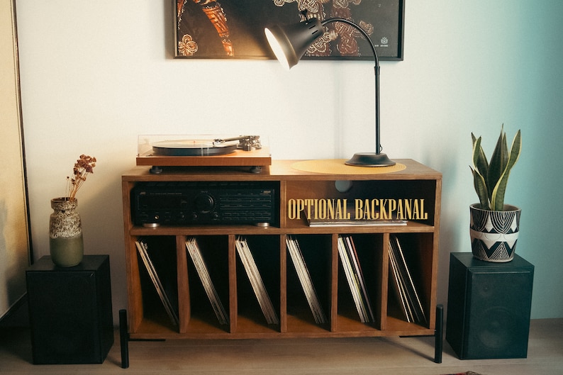 May include: A wooden record player stand with a turntable, amplifier, and shelves for vinyl records. The stand has a black lamp with a white shade and a plant in a black and white pot on top. The text "OPTIONAL BACKPANEL" is printed on the stand.