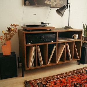 May include: A wooden record player stand with a turntable, amplifier, and shelves for vinyl records. The stand has a black lamp with a white shade and a vase with dried flowers on top. There is a rug on the floor in front of the stand.