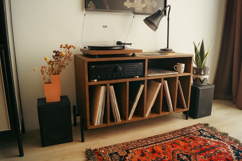 May include: A wooden record player stand with a turntable, amplifier, and shelves for vinyl records. The stand has a black lamp with a white shade and a vase with dried flowers on top. There is a rug on the floor in front of the stand.