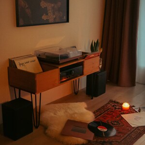 May include: A wooden record player stand with a turntable and a stack of vinyl records. The stand has black metal legs and is sitting on a light wood floor. There is a white furry rug and a red rug with a patterned design on the floor. A lit candle is on the red rug.