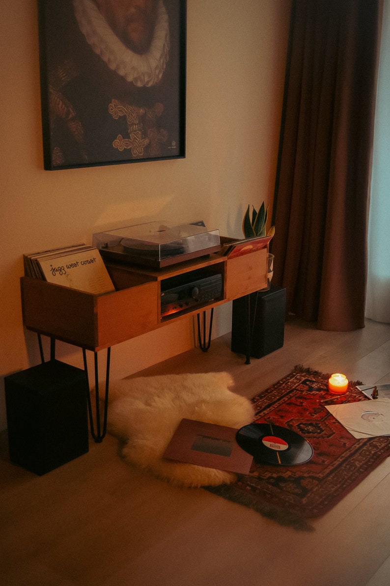 May include: A wooden record player stand with a turntable and a stack of vinyl records. The stand has black metal legs and is sitting on a light wood floor. There is a white furry rug and a red rug with a patterned design on the floor. A lit candle is on the red rug.