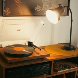 May include: A vintage-style record player with a black vinyl record on the turntable. The record player is on a wooden shelf with a lamp and a plant.