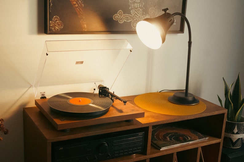 May include: A vintage-style record player with a black vinyl record on the turntable. The record player is on a wooden shelf with a lamp and a plant.