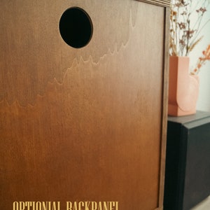 May include: A wooden cabinet with a brown finish and a round hole in the back panel. The text "OPTIONAL BACKPANEL" is printed on the cabinet.