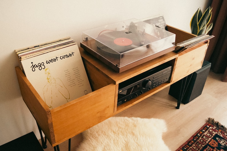 May include: A wooden record player stand with two compartments for storing vinyl records. The stand has a turntable with a clear lid and a black amplifier. The stand is on a light wood floor with a white rug and a small green plant.
