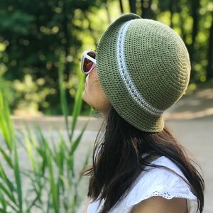 Peut inclure: Un chapeau en crochet vert en forme de seau avec une bordure blanche. Le chapeau est porté par une personne assise sur un chemin avec des plantes vertes en arrière-plan.
