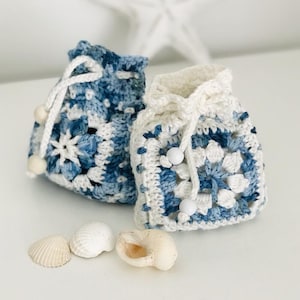 May include: Two crocheted drawstring bags with blue and white patterns. The bags are made with a granny square stitch and have white wooden beads on the drawstrings. The bags are sitting on a white surface with seashells.