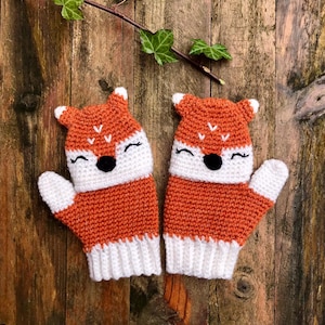 Puede incluir: Un par de guantes de punto de zorro naranja y blanco con ojos negros y sonrisas blancas. Los guantes están sobre un fondo de madera.