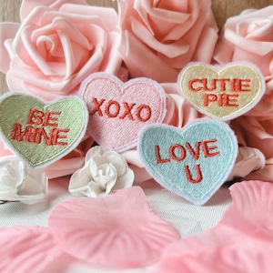 May include: Four heart-shaped embroidered patches with different messages: "Be Mine", "XOXO", "Cutie Pie", and "Love U". The patches are on a white background with pink rose petals.